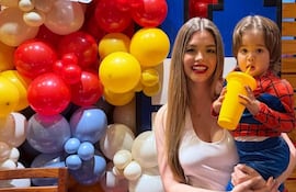 Mujer con camiseta blanca sonríe mientras sostiene a un niño en traje de Spider-Man, rodeados de globos coloridos.