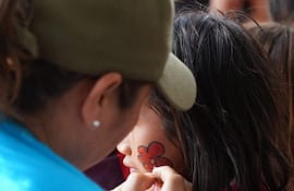 Joven con cabello largo y camiseta rosa recibe pintura en la mejilla de una persona con gorra y camiseta azul en evento al aire libre.
