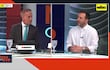 Ceres Escobar en traje y corbata turquesa, mientras otro hombre con barba escucha atentamente en un set de televisión.