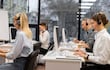 Tres mujeres y dos hombres en oficina moderna, usando auriculares; luz natural y elementos de trabajo visibles en escritorios.