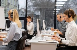 Tres mujeres y dos hombres en oficina moderna, usando auriculares; luz natural y elementos de trabajo visibles en escritorios.