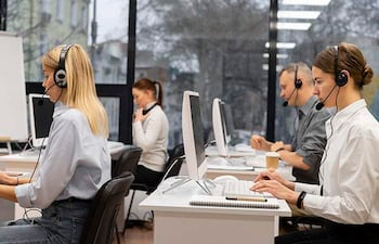 Tres mujeres y dos hombres en oficina moderna, usando auriculares; luz natural y elementos de trabajo visibles en escritorios.