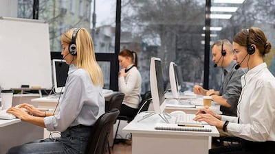 Tres mujeres y dos hombres en oficina moderna, usando auriculares; luz natural y elementos de trabajo visibles en escritorios.