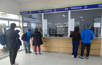 Cinco personas en la farmacia del IPS, entre ellas una mujer con chaqueta negra y un hombre con camiseta azul, esperan atención.
