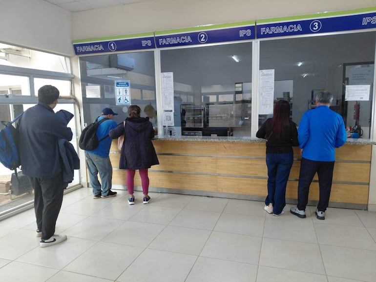 Cinco personas en la farmacia del IPS, entre ellas una mujer con chaqueta negra y un hombre con camiseta azul, esperan atención.