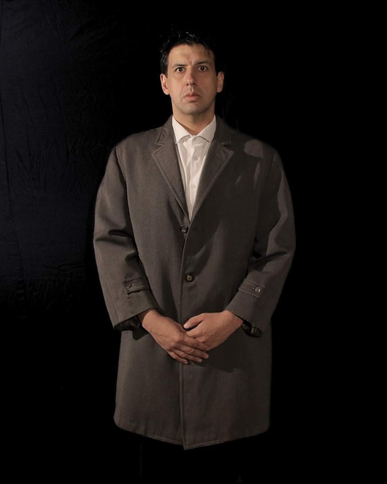 Hombre de pie con abrigo gris y camisa blanca, con expresión seria, en un fondo negro.