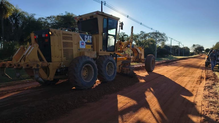 Máquina niveladora Caterpillar trabaja en camino de tierra, rodeada de árboles y postes eléctricos en un ambiente soleado.