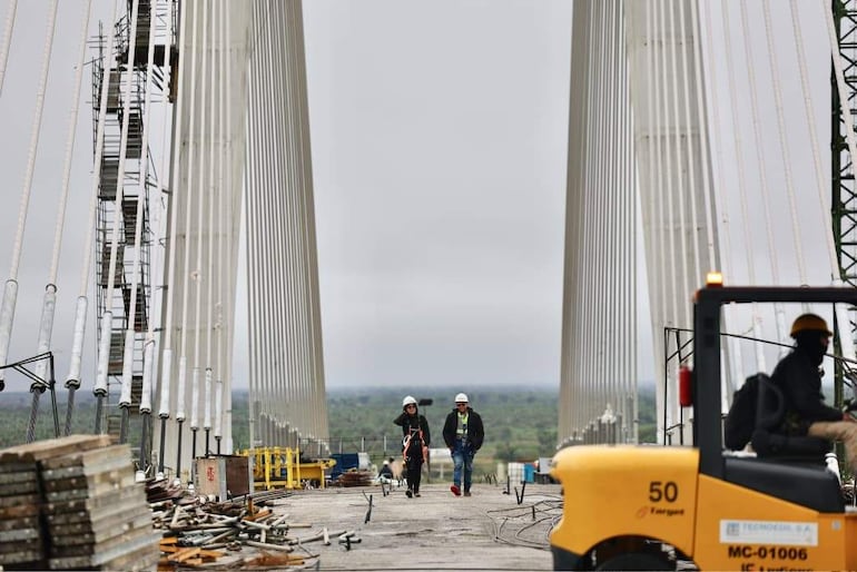 Dos trabajadores con cascos blancos, uno con chaqueta negra y otro con camiseta verde, supervisan la construcción bajo un cielo nublado.