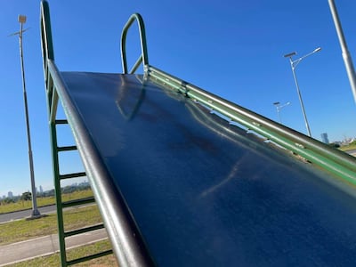 Resbaladilla de metal verde en costanera vacía bajo cielo azul, sin personas ni actividad recreativa visible.