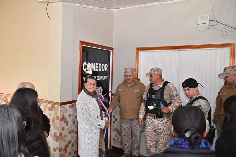 Mujer mayor con abrigo claro sosteniendo un lazo, rodeada de hombres en uniformes militares, observadores tras ella.
