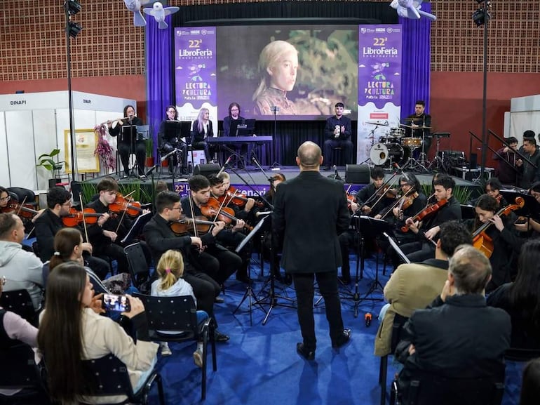 Grupo de músicos vestidos de negro tocando violines en un escenario decorado, mientras el director de orquesta los guía desde atrás.
