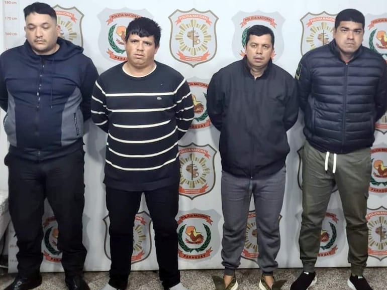 Cuatro hombres de pie en un ambiente institucional, con vestimenta oscura, expresiones neutrales, alineados frente a emblemas policiales.
