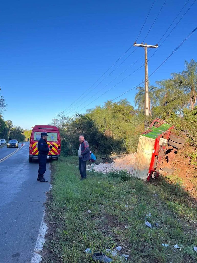 Policía en uniforme habla con hombre mayor en la orilla de una carretera, mientras un camión volcado y vegetación rodean la escena.