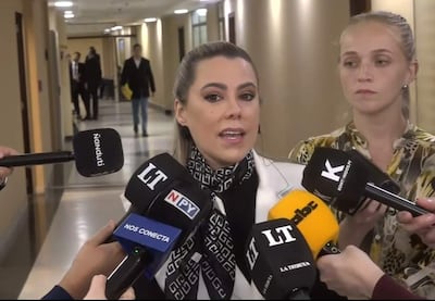 Lizarella Valiente con blusa blanca y cuello negro, visiblemente triste, mientras habla a periodistas en un pasillo formal.