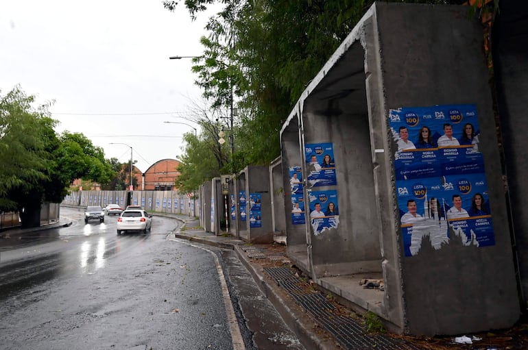 Carteles políticos en la calle Gral Santos, bajo la lluvia, con colores azul y blanco y sin personas visibles.