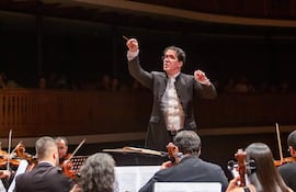 Director de orquesta en chaleco blanco y chaqueta oscura, gesticulando con baqueta entre músicos tocando violines en un auditorio elegante.