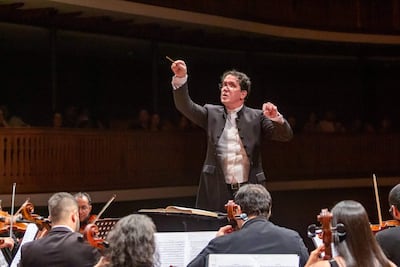 Director de orquesta en chaleco blanco y chaqueta oscura, gesticulando con baqueta entre músicos tocando violines en un auditorio elegante.