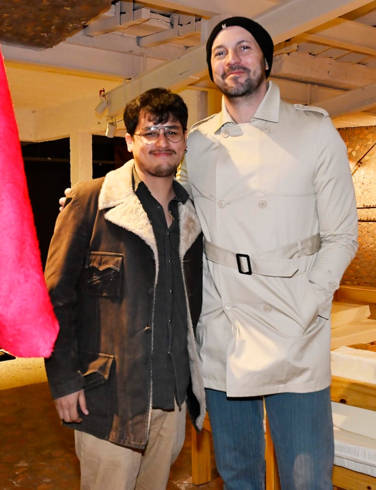 Dos hombres sonrientes, uno en chaqueta oscura y otro en abrigo claro, posan juntos en un entorno iluminado.