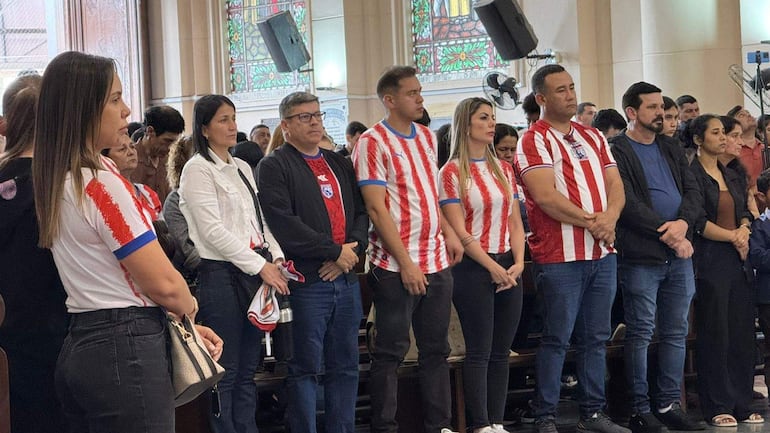 Grupo de personas con camisetas rojas y blancas en ceremonia, algunos escuchando atentamente en un entorno solemne con vitrales.
