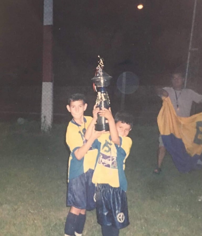 Dos niños con camisetas amarillas sonríen mientras sostienen un trofeo en un ambiente festivo.