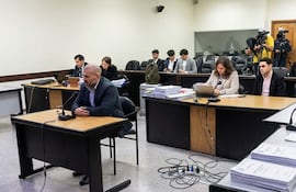 Hugo Ramírez, con abrigo oscuro y barba, habla al micrófono en una sala de tribunal; una mujer toma notas a su lado.