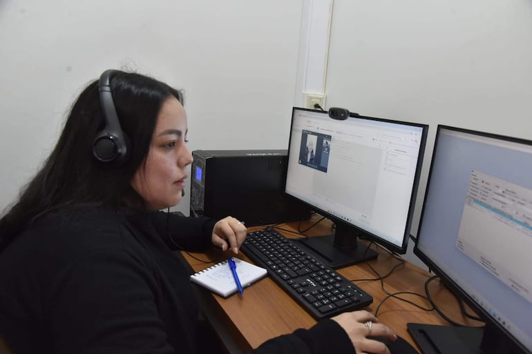 Mujer con auriculares frente a dos computadoras en oficina sencilla, tomando notas mientras realiza videoconferencia.