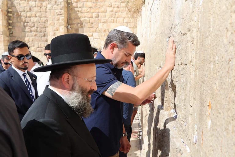 Seis hombres, uno con sombrero negro y otro con yarmulke, tocan el Muro de las Lamentaciones en actitud de oración.