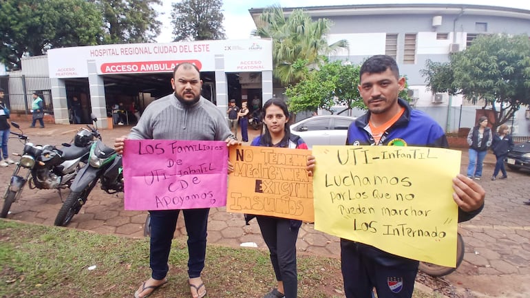 Tres personas están de pie sosteniendo carteles de protesta frente al Hospital Regional, luciendo camisetas de diferentes colores.
