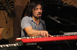 Rolfi Gómez en camisa azul, concentrado al piano negro, rodeado de arte abstracto en un ambiente acogedor.