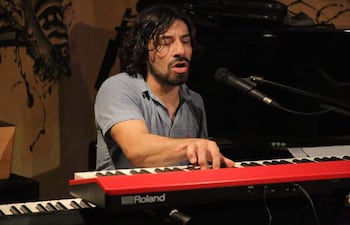 Rolfi Gómez en camisa azul, concentrado al piano negro, rodeado de arte abstracto en un ambiente acogedor.