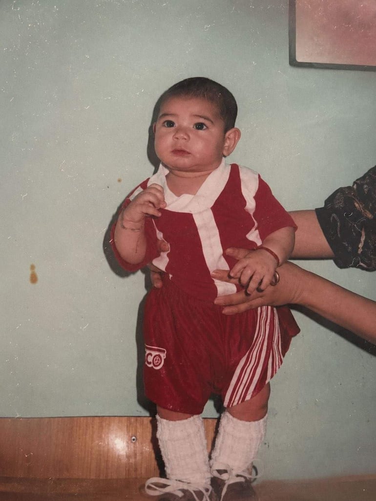 Niño con camiseta roja y pantalones cortos, sostenido por un adulto, mirando a la cámara con curiosidad.