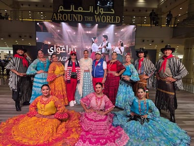 Grupo de diez personas en trajes folclóricos coloridos posan con alegría frente a un cartel que dice 'AROUND THE WORLD'.