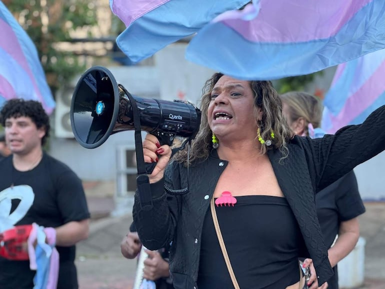 Persona con megáfono y cabello rizado, rodeada de manifestantes con banderas rosa y celeste en un acto de protesta.
