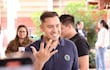 Hombre sonriente con camiseta oscura y logo verde muestra su dedo con tinta en un centro de votación.
