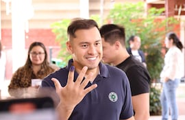 Hombre sonriente con camiseta oscura y logo verde muestra su dedo con tinta en un centro de votación.