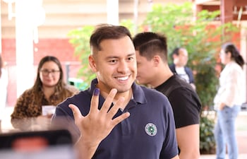 Hombre sonriente con camiseta oscura y logo verde muestra su dedo con tinta en un centro de votación.