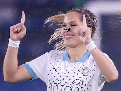 Jugadora del Club Libertad con camiseta blanca y azul, levantando el dedo índice en gesto de celebración en fondo deportivo azul.