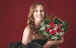 Mujer agachada con vestido negro y tacones altos, sonriente, sosteniendo un ramo de flores en un fondo rojo.