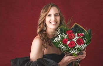 Mujer agachada con vestido negro y tacones altos, sonriente, sosteniendo un ramo de flores en un fondo rojo.