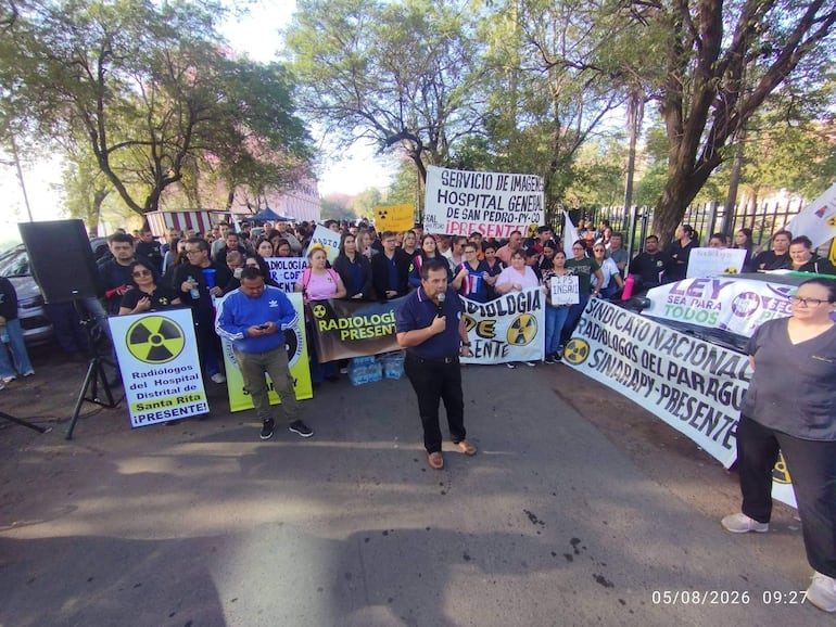 Gran grupo de radiólogos con camisetas profesionales y pancartas, un hombre habla por micrófono, ambiente de protesta.