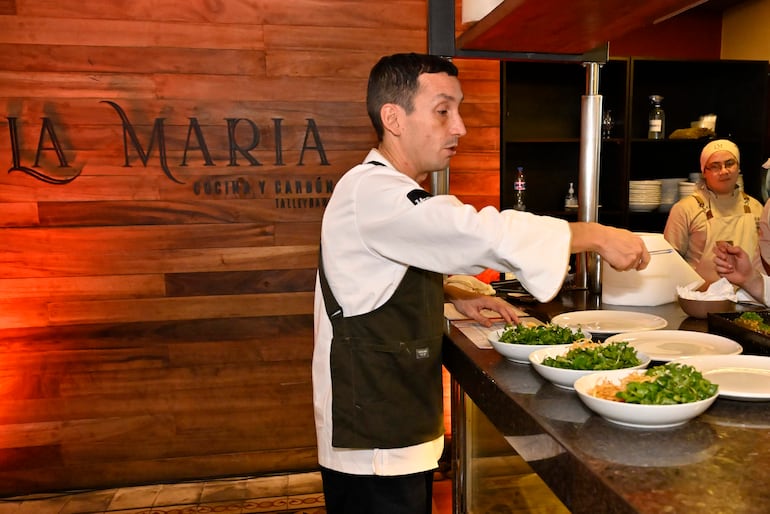 Hombre vestido de chef sirve comida en platos mientras una mujer con delantal observa en la cocina de Talleyrand.