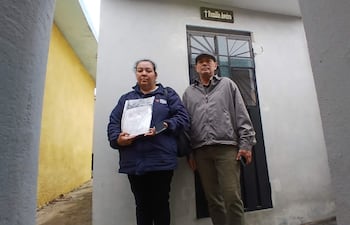 Felicia Ramona Benítez Aguilera con chaqueta azul, sosteniendo un documento, mientras un hombre con sombrero gris se encuentra a su lado.