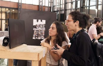Dos mujeres en blusa clara y falda larga, y camiseta negra con gafas, observan una pantalla en un ambiente luminoso.