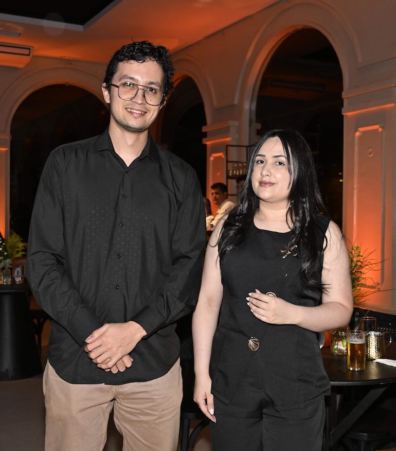 Hombre con camisa negra y pantalones claros sonríe junto a mujer en conjunto oscuro, ambos en un ambiente festivo y elegante.