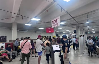 Hombre con gorra y camiseta blanca dialoga con otro en camiseta roja, rodeados de personas en un ambiente interior de atención al público.