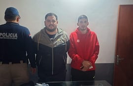 Tres hombres en oficina; uno de espaldas con camiseta de 'POLICÍA ANTINARCÓTICOS', dos de pie vistiendo sudadera roja y chaqueta negra.