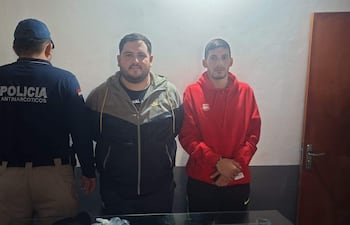 Tres hombres en oficina; uno de espaldas con camiseta de 'POLICÍA ANTINARCÓTICOS', dos de pie vistiendo sudadera roja y chaqueta negra.