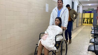Mujer en silla de ruedas cubierta con manta blanca, acompañada por hombre de chaqueta blanca, mientras otros pacientes esperan en el pasillo.