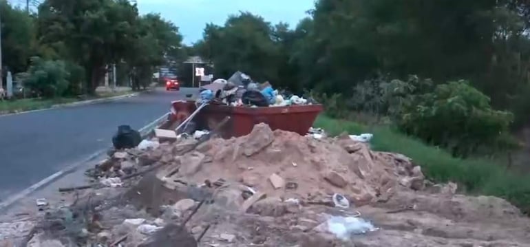 Montón de basura y escombros en la vereda de una carretera, sin personas visibles y un vehículo al fondo.