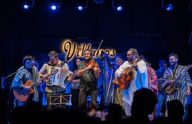 Seis músicos en el escenario: un guitarrista con poncho, un acordeonista, un violinista, una cantante y otros con guitarra y saxofón.